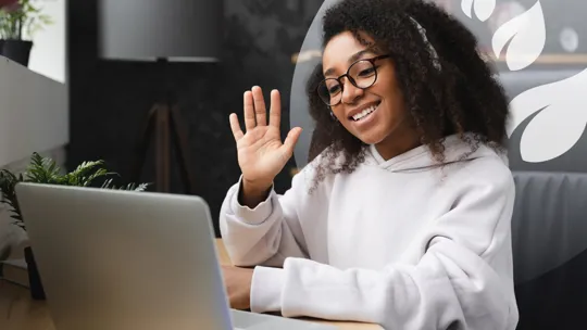A young person with glasses and headphones, smiling and waving at a laptop screen, engaged in an online video call.