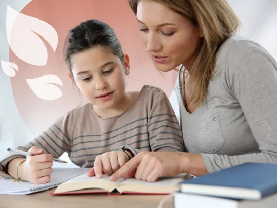 A homeschooling mother and daughter sat at a desk reading through a book. The girl is holding a pen with a notepad. The mother is helping the child homeschool.