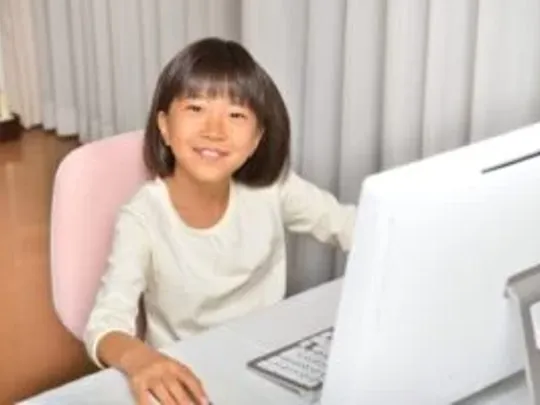 A photograph of a Primary-age girl with black hair sitting at a desktop computer and smiling at the camera.