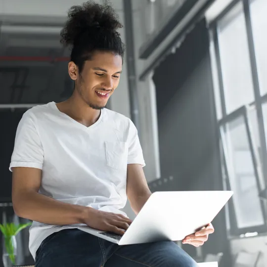 A young man in a white t-shirt and hair in a bun smiles while working on a laptop by a large, sunny window in a modern office, representing flexible online learning.