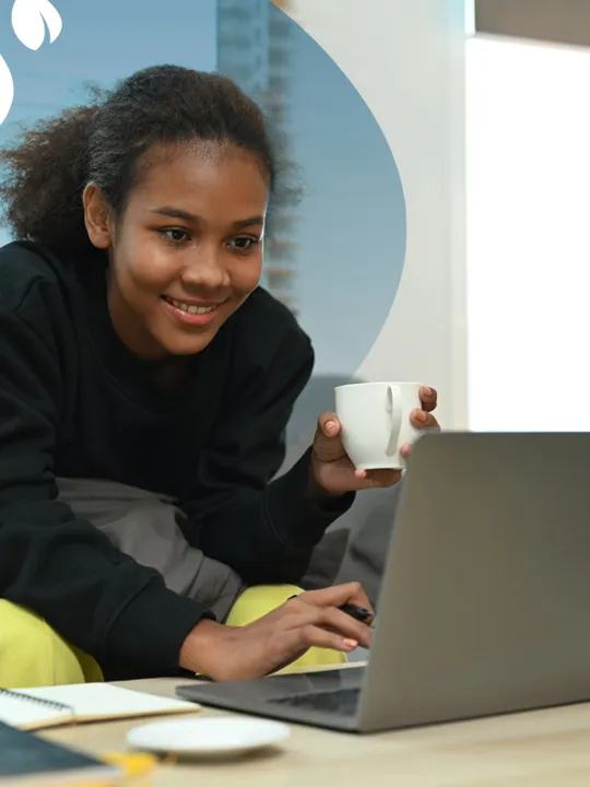 Smiling student with a mug in hand using a laptop at home, seated on a sofa with notebooks on the table.