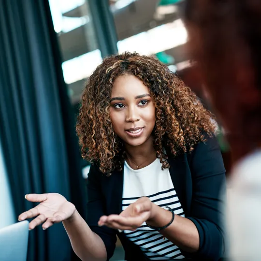 A young woman with a warm smile stands between two classmates, engaged in a conversation outdoors in a modern, garden-like setting.