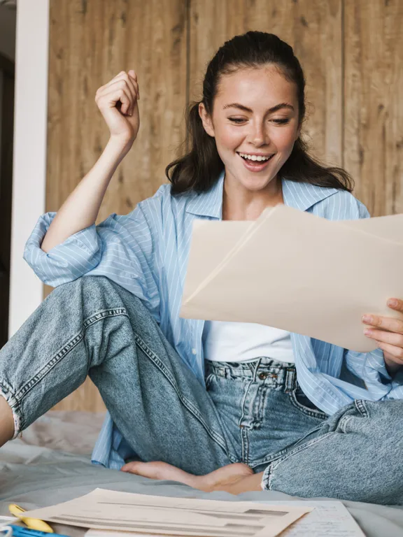 A delighted Wolsey Hall student sits on her bed, excitedly reviewing documents and celebrating her achievement as a Cambridge Learner Award winner.