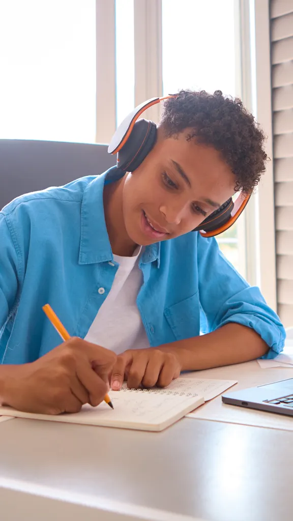 A young student wearing headphones diligently writes in a notebook at their desk, using a laptop, likely preparing for a mock exam.