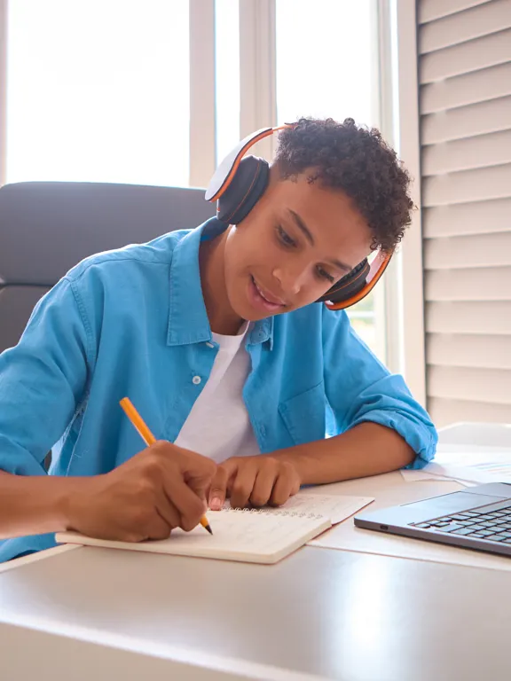 A young student wearing headphones diligently writes in a notebook at their desk, using a laptop, likely preparing for a mock exam.