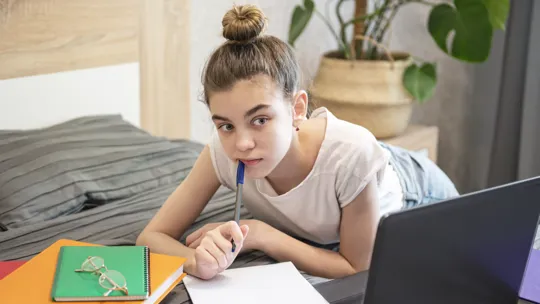 Teen girl lying on bed, thoughtfully holding a pen while studying with laptop and folders.