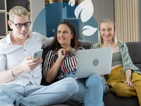 Three people sitting on a sofa smiling while looking at a laptop and phone together