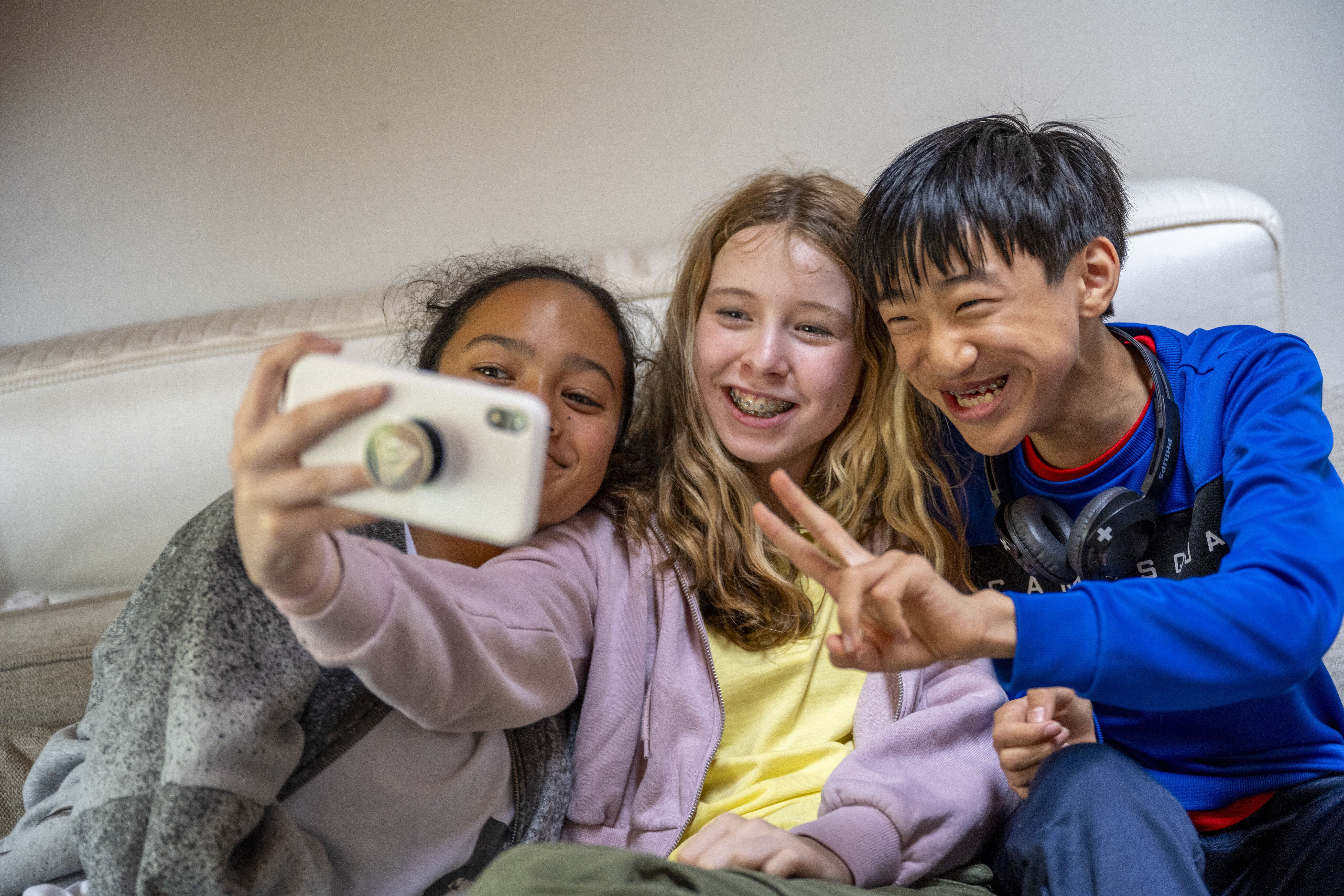 Three smiling teenagers taking a selfie together, sitting close on a sofa.