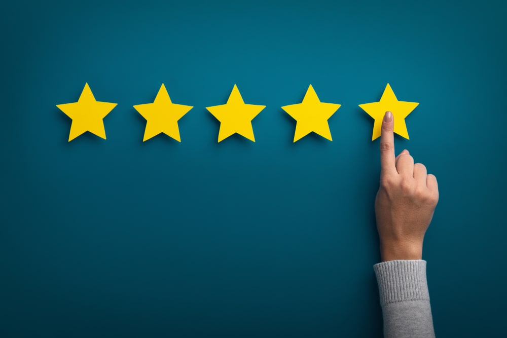 A hand pointing to the fifth yellow star in a row of five, symbolising a top 5-star rating or excellent review, against a teal blue background.