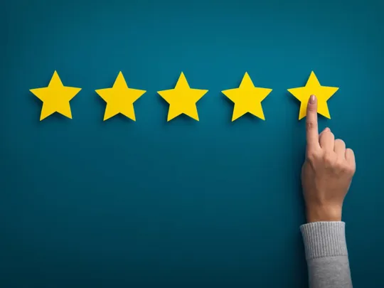A hand pointing to the fifth yellow star in a row of five, symbolising a top 5-star rating or excellent review, against a teal blue background.