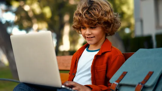 Young learner focused on a laptop while sitting on a bench in a leafy outdoor setting.