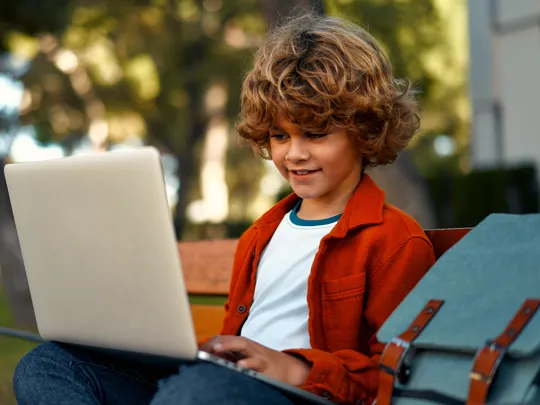 Young learner focused on a laptop while sitting on a bench in a leafy outdoor setting.