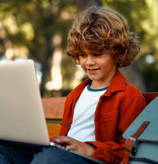 Young learner focused on a laptop while sitting on a bench in a leafy outdoor setting.