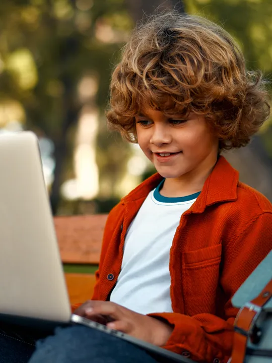 Young learner focused on a laptop while sitting on a bench in a leafy outdoor setting.