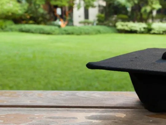 Black graduation cap resting on a wooden bench in a peaceful garden setting.