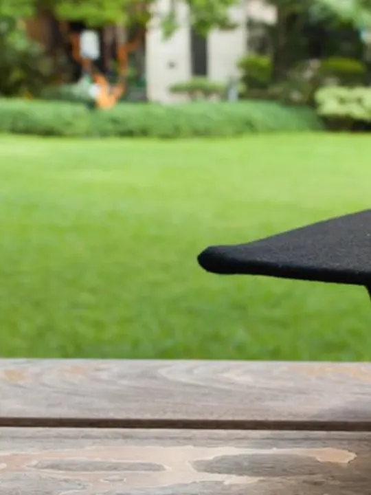 Black graduation cap resting on a wooden bench in a peaceful garden setting.