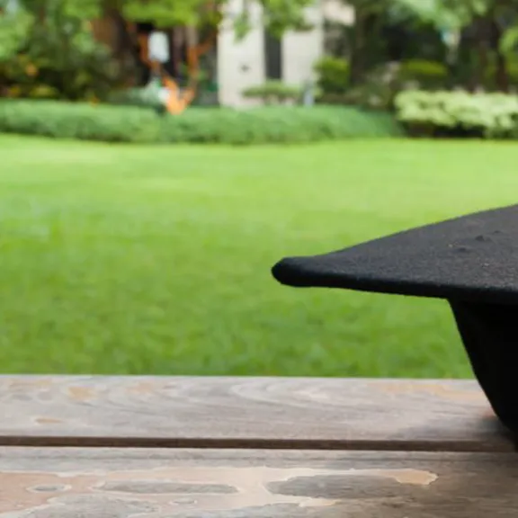 Black graduation cap resting on a wooden bench in a peaceful garden setting.