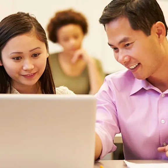 A male teacher or tutor wearing a pink shirt is smiling while leaning in to help a female student look at content on her laptop screen in a classroom setting. Another student is blurred in the background.