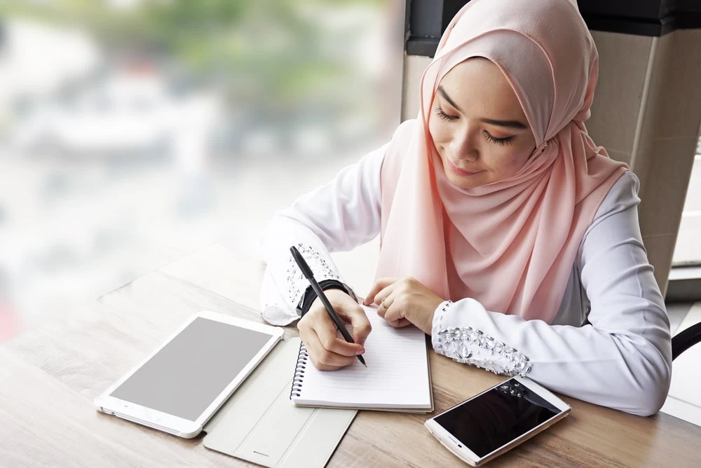 A female student studying, using her tablet and making notes in her notepad. 