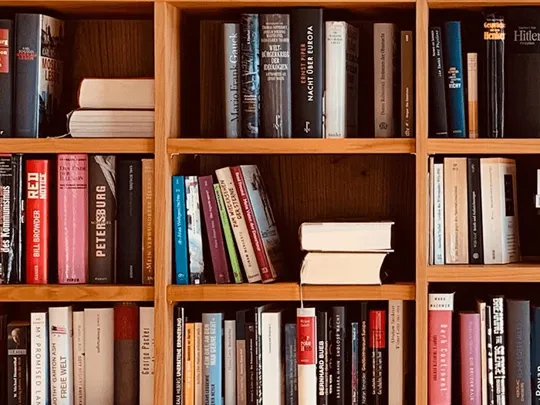 A close-up shot of several wooden bookshelves filled with various books. Some books are standing upright, while others are stacked horizontally. The spines of the books are different colors and have different titles, although they are not clearly readable.