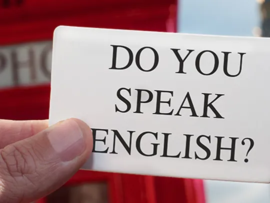 Close-up of a hand holding a card that reads ‘Do you speak English?’ with a red telephone box in the background.