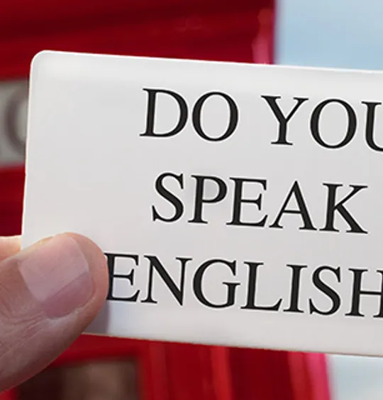 Close-up of a hand holding a card that reads ‘Do you speak English?’ with a red telephone box in the background.