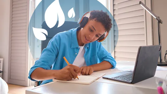 A young person wearing headphones, writing in a notebook at a desk with a laptop and school supplies.