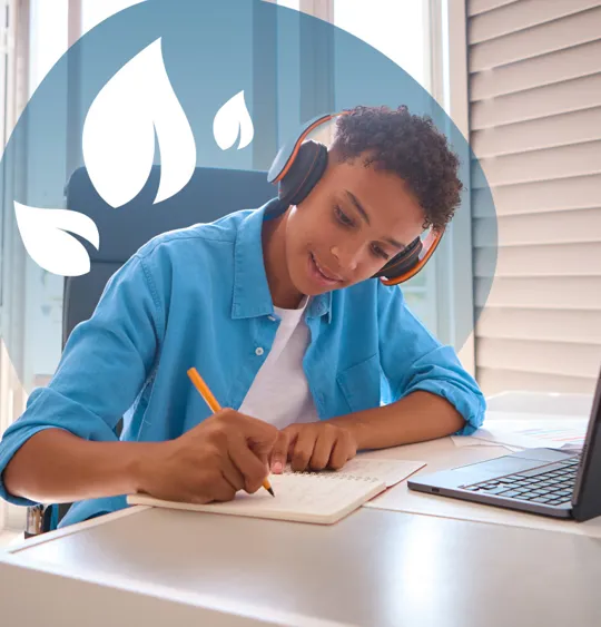 A young person wearing headphones, writing in a notebook at a desk with a laptop and school supplies.