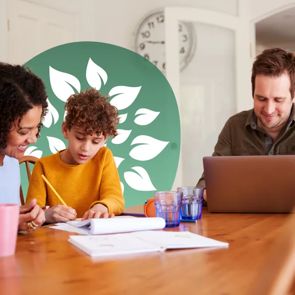 Mother helping child with work at a table while father works on a laptop nearby