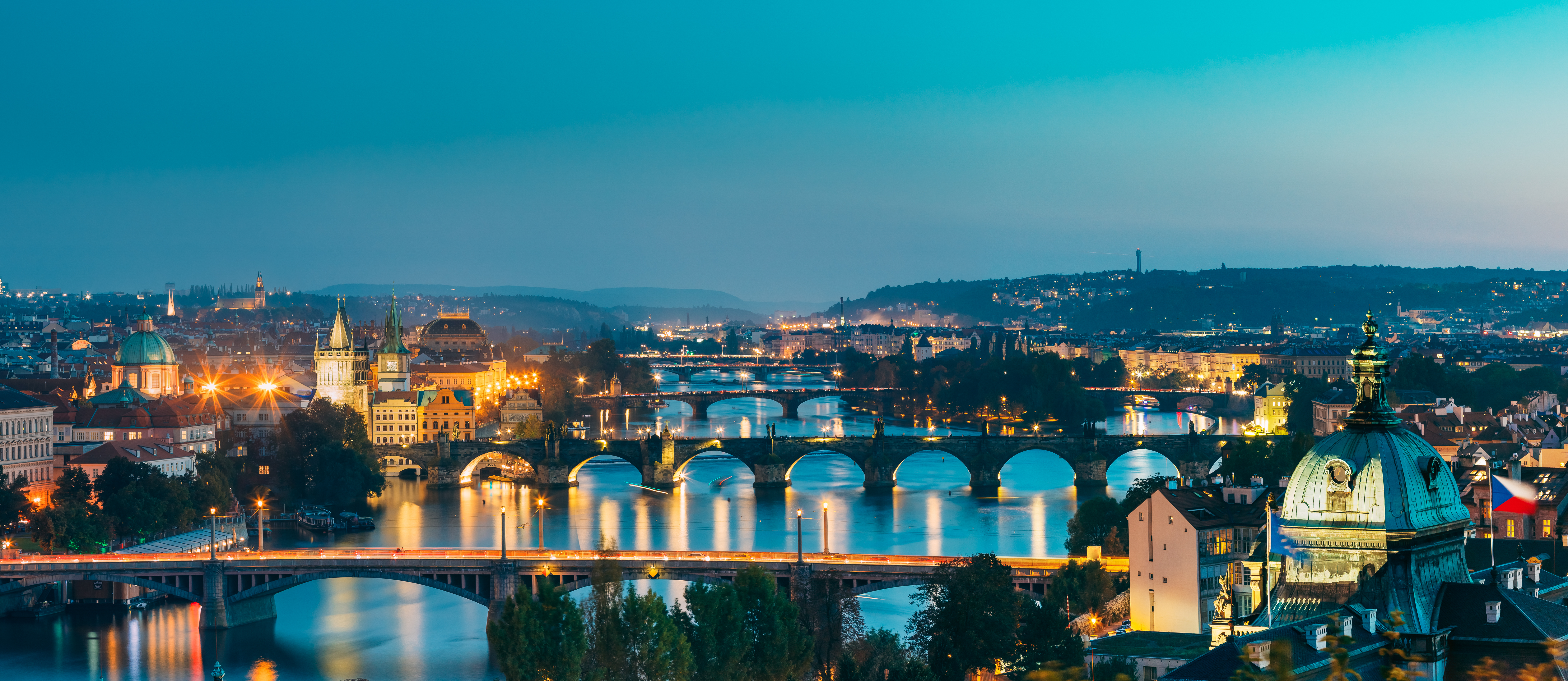 Panoramic twilight view of Prague with iconic bridges and historic architecture lit up.
