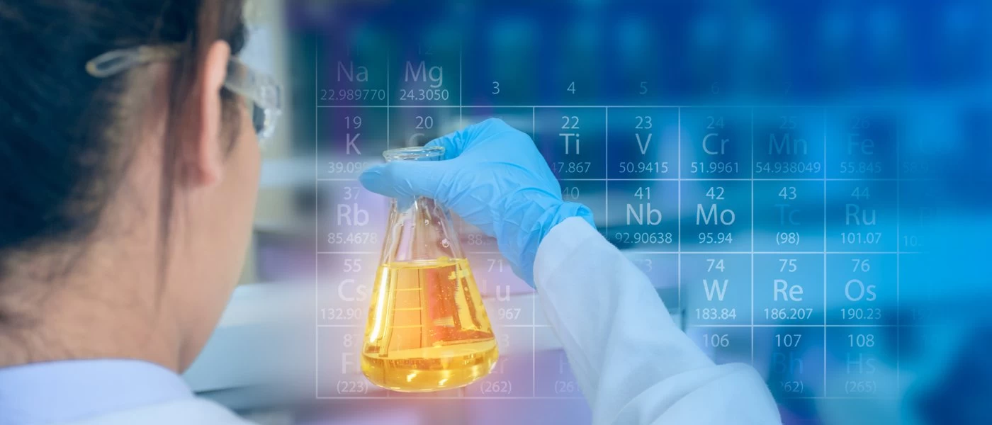 A scientist looking at a science beaker with orange liquid in it. A semi transparent periodic table is laid over the image.
