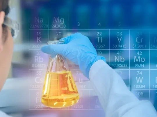 A scientist looking at a science beaker with orange liquid in it. A semi transparent periodic table is laid over the image.