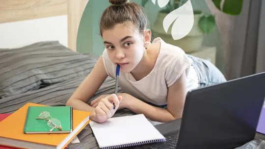 Teen girl lying on bed, thoughtfully holding a pen while studying with laptop and folders.