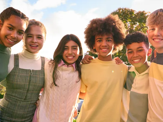 Group of six smiling children standing close together outdoors on a sunny day.