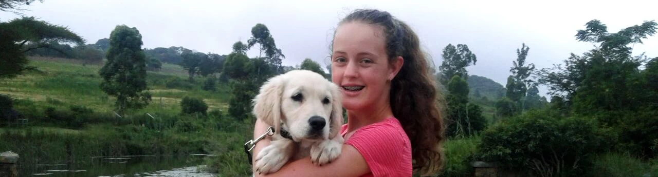 A young woman smiling and holding a light-coloured puppy, standing in a natural landscape with trees and water in the background.