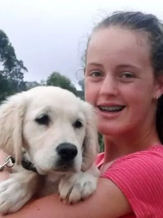 A young woman smiling and holding a light-coloured puppy, standing in a natural landscape with trees and water in the background.