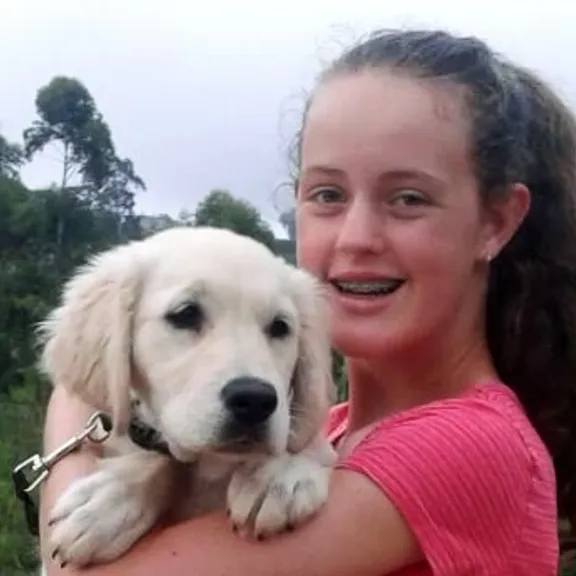 A young woman smiling and holding a light-coloured puppy, standing in a natural landscape with trees and water in the background.