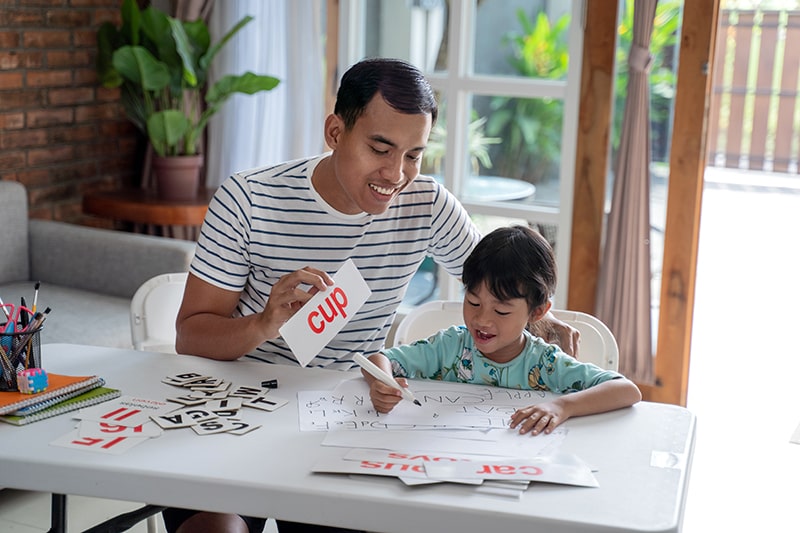 Parent supporting child with phonics activity at home, using flashcards and writing practice on a table