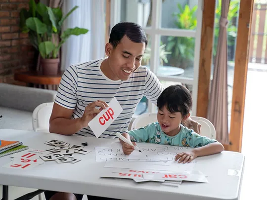 Parent supporting child with phonics activity at home, using flashcards and writing practice on a table