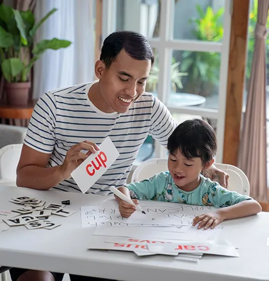 Parent supporting child with phonics activity at home, using flashcards and writing practice on a table