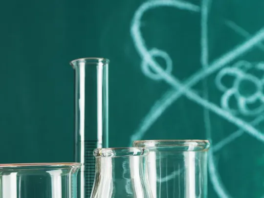 Beakers and test tubes in front of a chalkboard with an atom diagram and colourful pens.