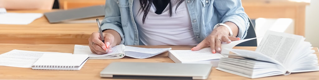 Learner writing notes with study materials and laptop on a wooden desk.