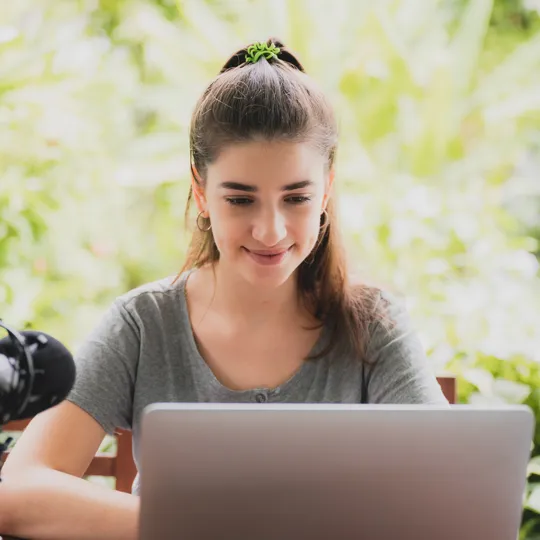 A young woman with her hair in a ponytail smiles while working on a laptop with a professional microphone in an outdoor, lush green setting, representing flexible online learning.