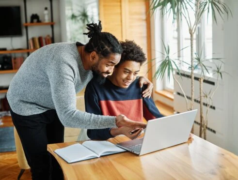 A homeschooling dad and son working together on a laptop at the kitchen table.
