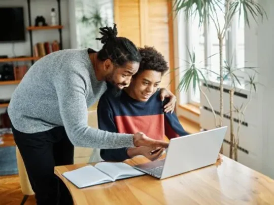 A homeschooling dad and son working together on a laptop at the kitchen table.