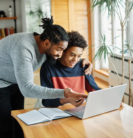 Father smiling and guiding his teenage son through online studies on a laptop at home.