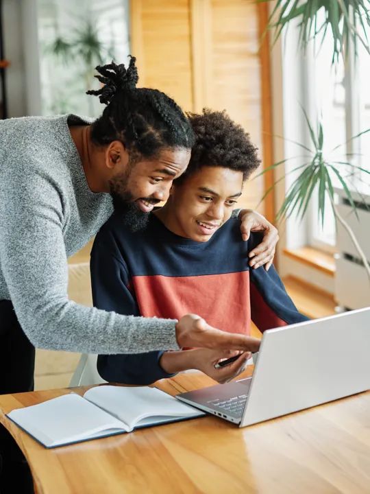 Father smiling and guiding his teenage son through online studies on a laptop at home.