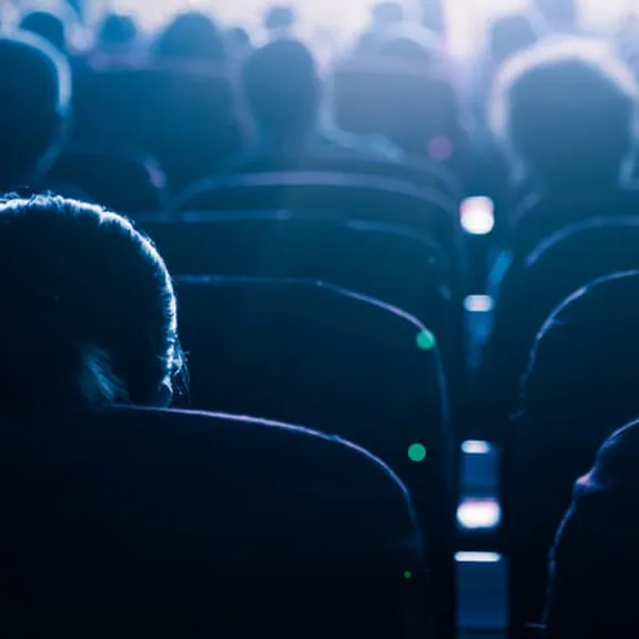A view from behind of people sitting in a dark cinema or theater, watching a screen. The light from the screen illuminates the tops of their heads and the seats.