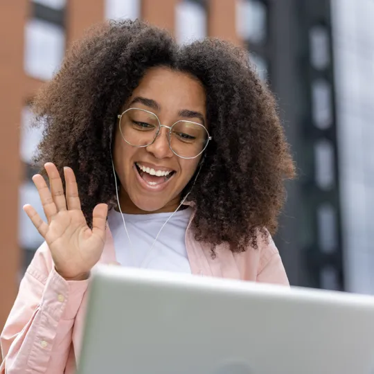 A young woman with curly hair and glasses smiles and waves during an outdoor video call on her laptop, representing flexible online learning.