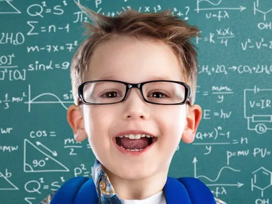 Smiling child wearing glasses and backpack, standing in front of chalkboard filled with maths and science formulas